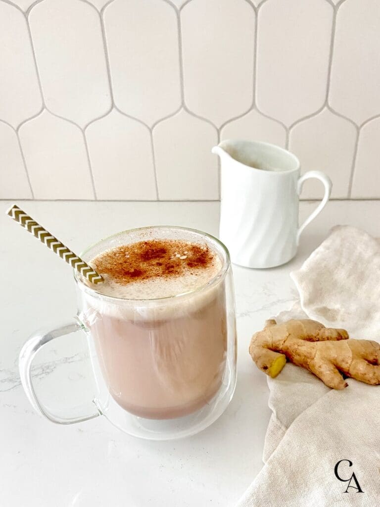 A clear glass mug with hot chocolate sprinkled with cinnamon, and a white ceramic pitcher next to a linen napkin on a white counter with white tile backsplash.