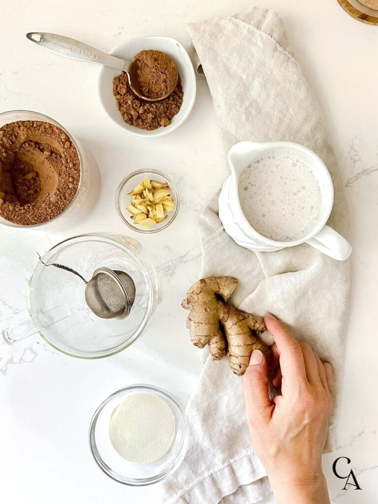 A hand holding ginger root next to a pitcher of almond milk, and cacao in a bowl.
