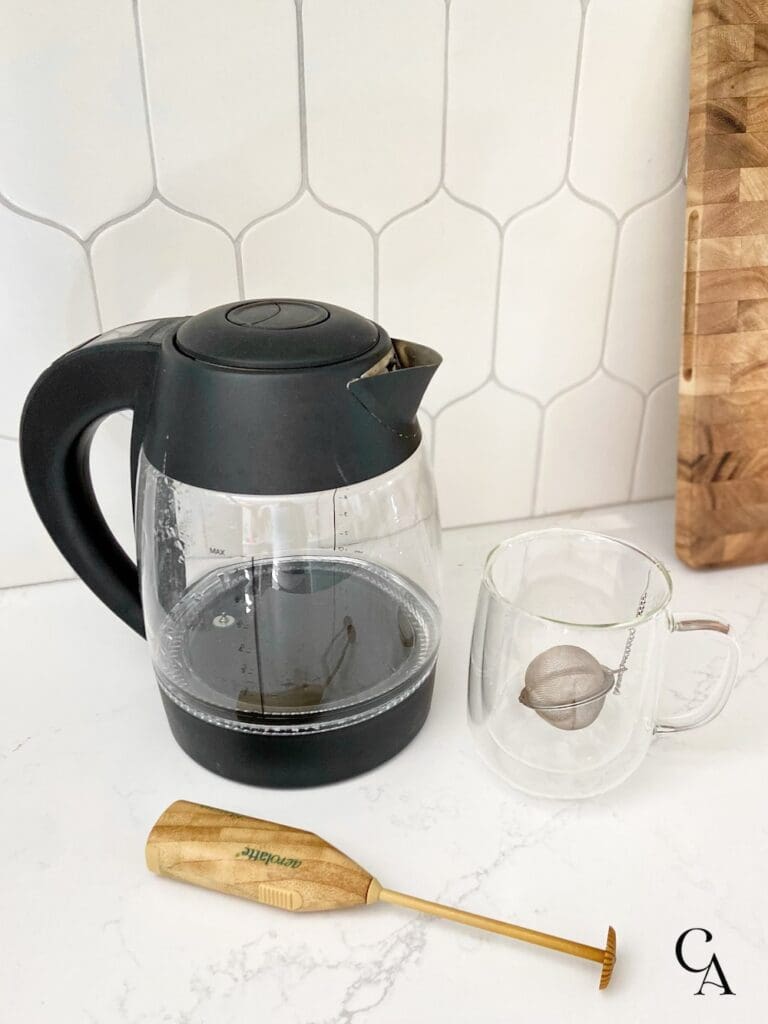 An electric kettle, clear glass hot chocolate mug and a latte frother on a white countertop.