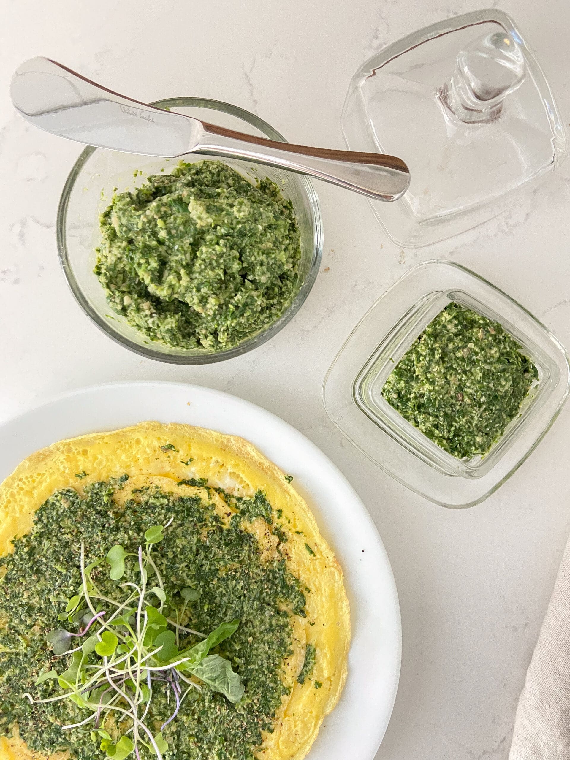 Open-faced omelet on a white place with basil pesto with walnuts spread on top, Prepared pesto in clear glass containers sitting on a white countertop next to the place.