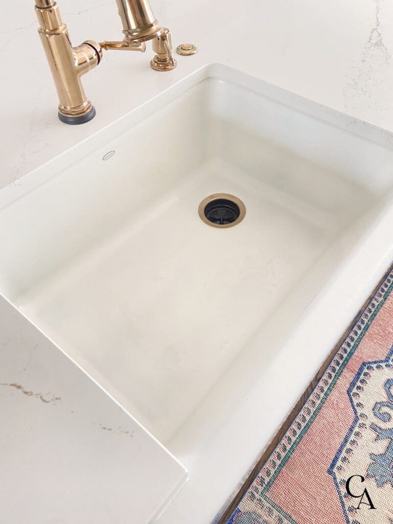 A clean, white, cast iron, farmhouse style kitchen sink with gold faucet.