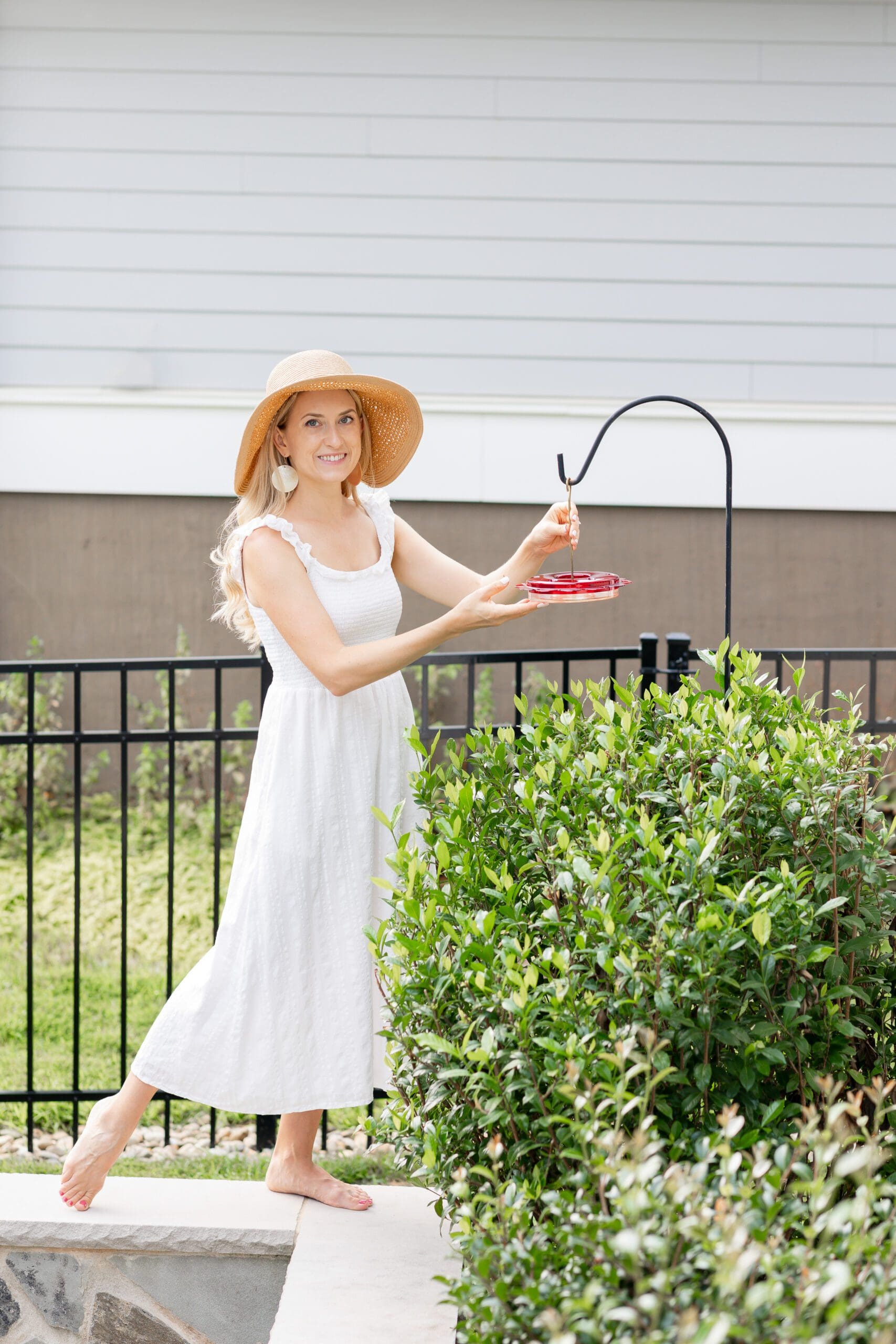 Woman in a white dress hanging a hummingbird feeder in a garden.