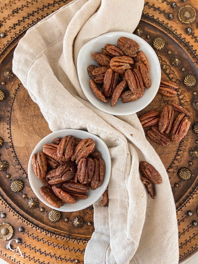 two small bowls of pumpkin spiced pecans with a linen napkin on a wood bread board.