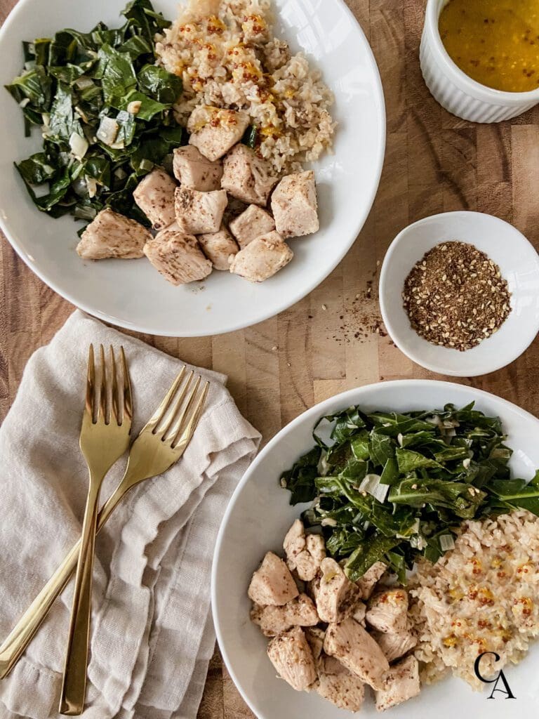 Two white bowls of greens and chicken with a linen napkin and two gold forks. A bowl of orange vinaigrette sits to the side.