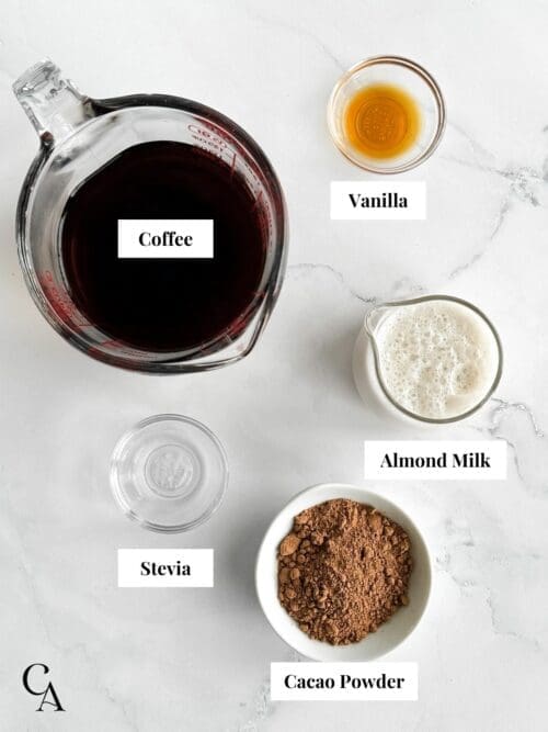 A measuring cup of coffee, a creamer of almond milk and pinch bowls of cacao powder, vanilla and stevia. 