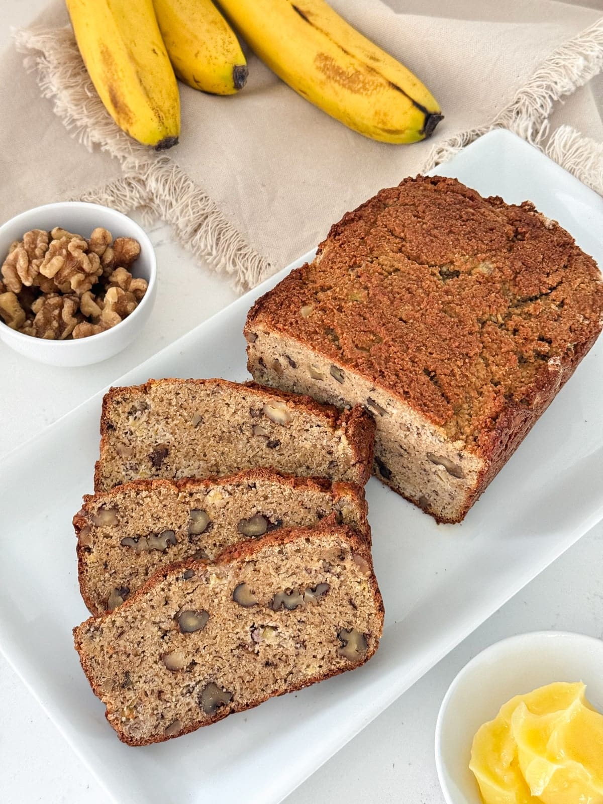 Three slices of gluten-free banana bread on a white plate, next to bowls of walnuts and ghee.