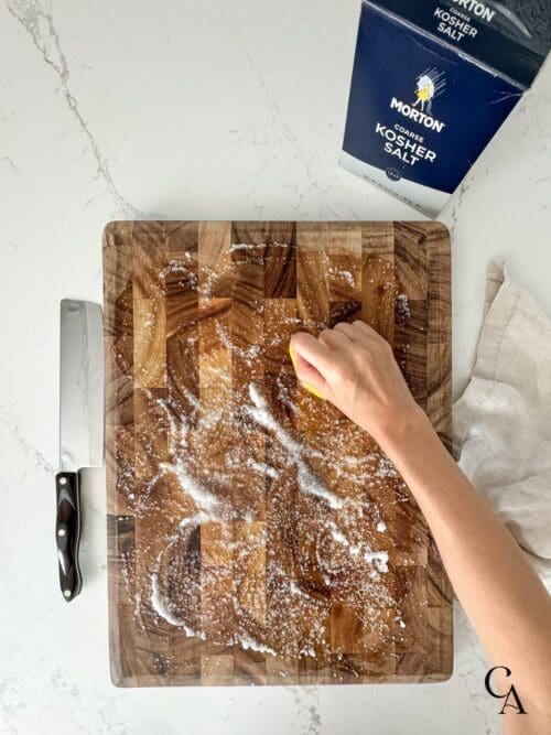 A woman's hand scrubbing a wood cutting board with lemon and salt.