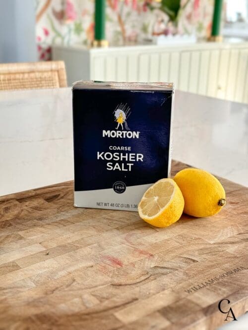 A wood cutting board with a box of kosher salt and a sliced lemon.