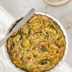 A breakfast casserole in a white tart dish with a silver knife and linen napkin.