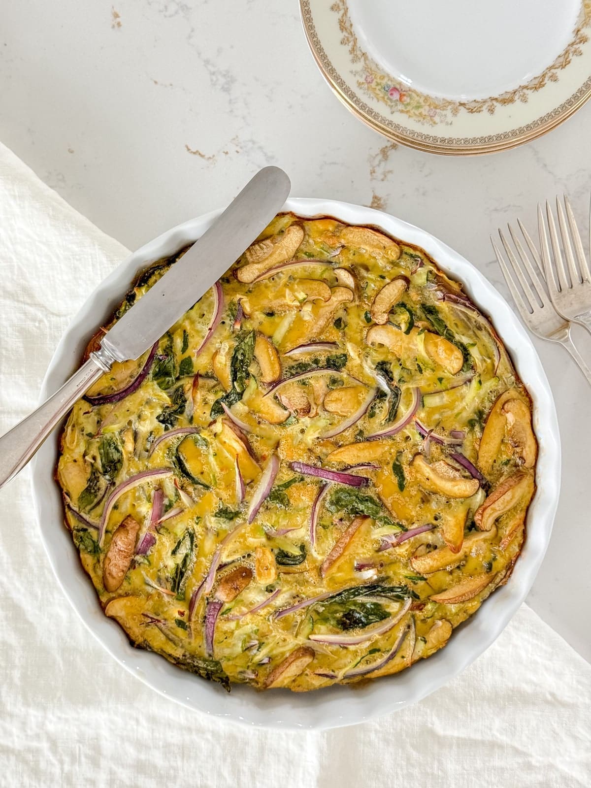 A breakfast casserole in a white tart dish with a silver knife and linen napkin.