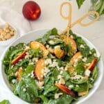 A spinach and nectarine salad in a large bowl with gold tongs.