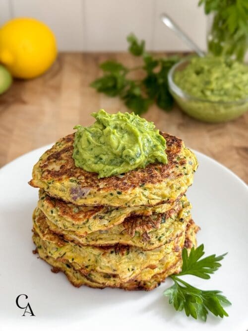 A stack of zucchini fritters on a white plate, topped with avocado gremolata.