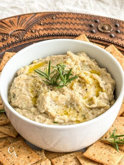 A bowl of white bean and artichoke dip with rosemary and crackers.