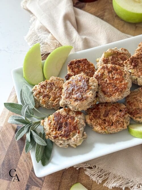 A platter of sausage patties with apple slices and fresh sage.