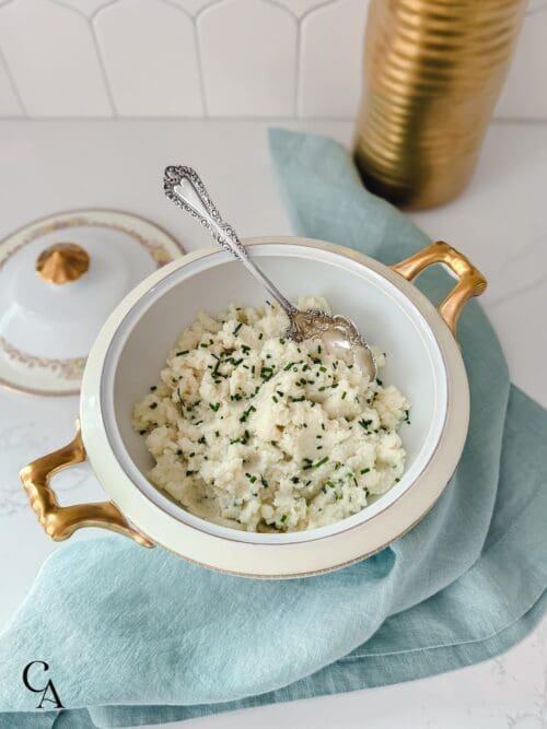 A bowl of creamy cauliflower mash with chives and a serving spoon.