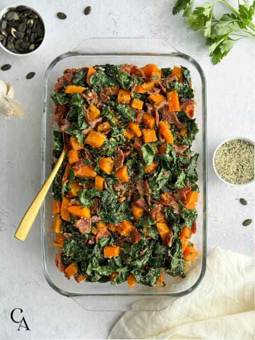 A casserole with butternut squash, kale, and turkey bacon with seeds on the side.