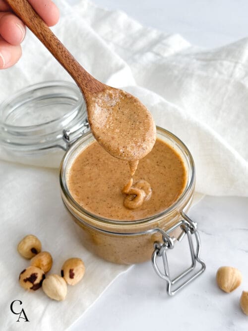 A hand holding a spoon over a jar of hazelnut butter.