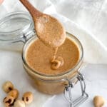 A glass jar of hazelnut butter with a wooden spoon on a white cloth.