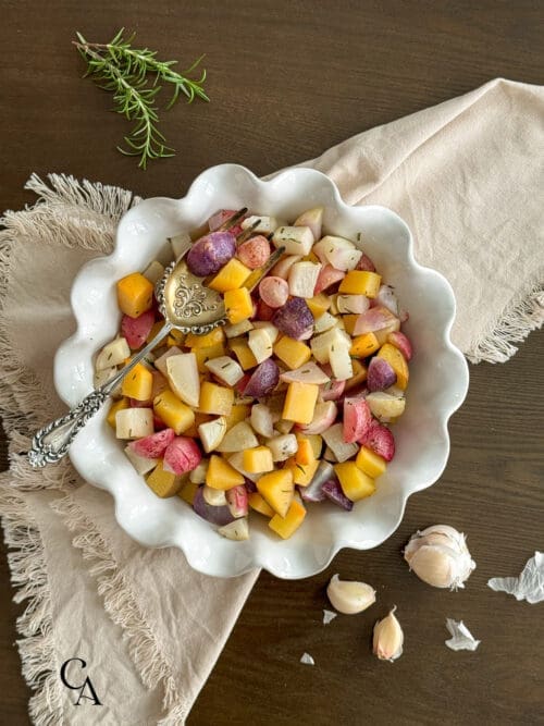 A dish of roasted root vegetables with rosemary and a silver serving spoon.