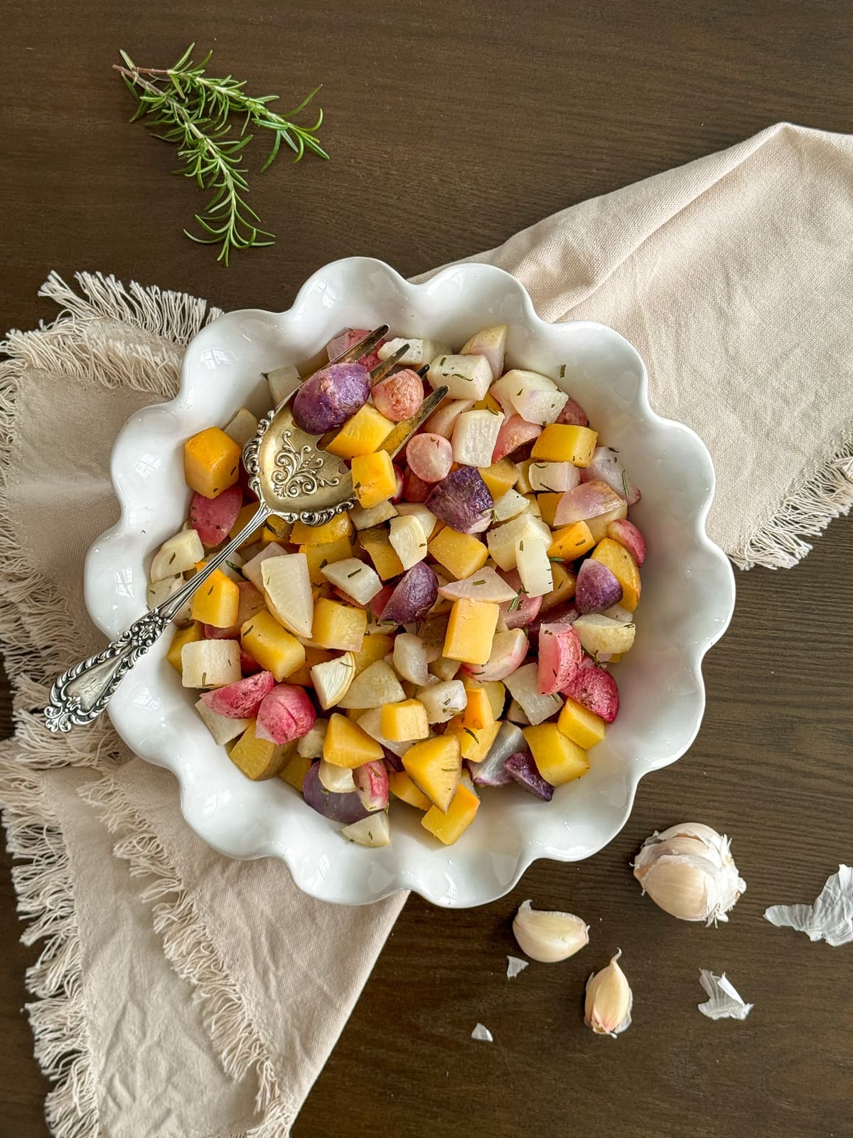 A dish of roasted root vegetables with rosemary and a silver serving spoon.