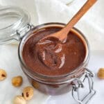 A container of chocolate hazelnut spread with cinnamon sticks and a small wooden spoon.