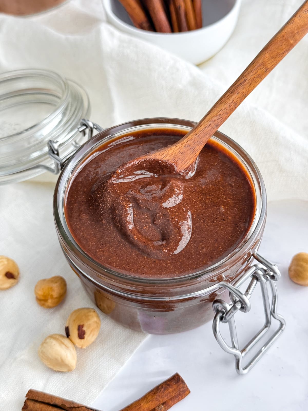 A container of chocolate hazelnut spread with cinnamon sticks and a small wooden spoon.