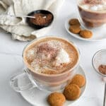 A tiramisu latte in a glass mug, on a white plate with small cookies.